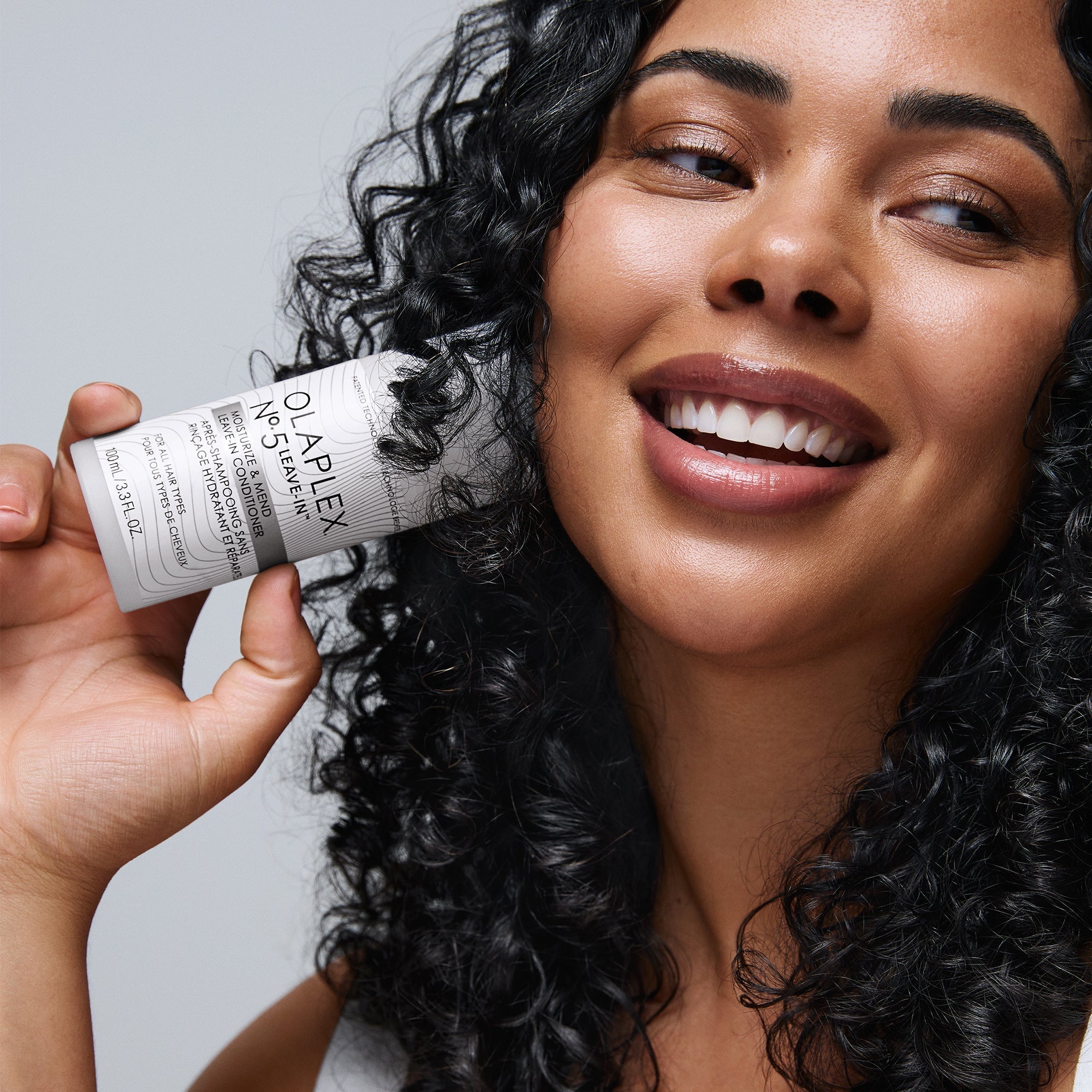 Woman with dark curly hair smiling and holding OLAPLEX No.5LEAVE-IN next to her face
