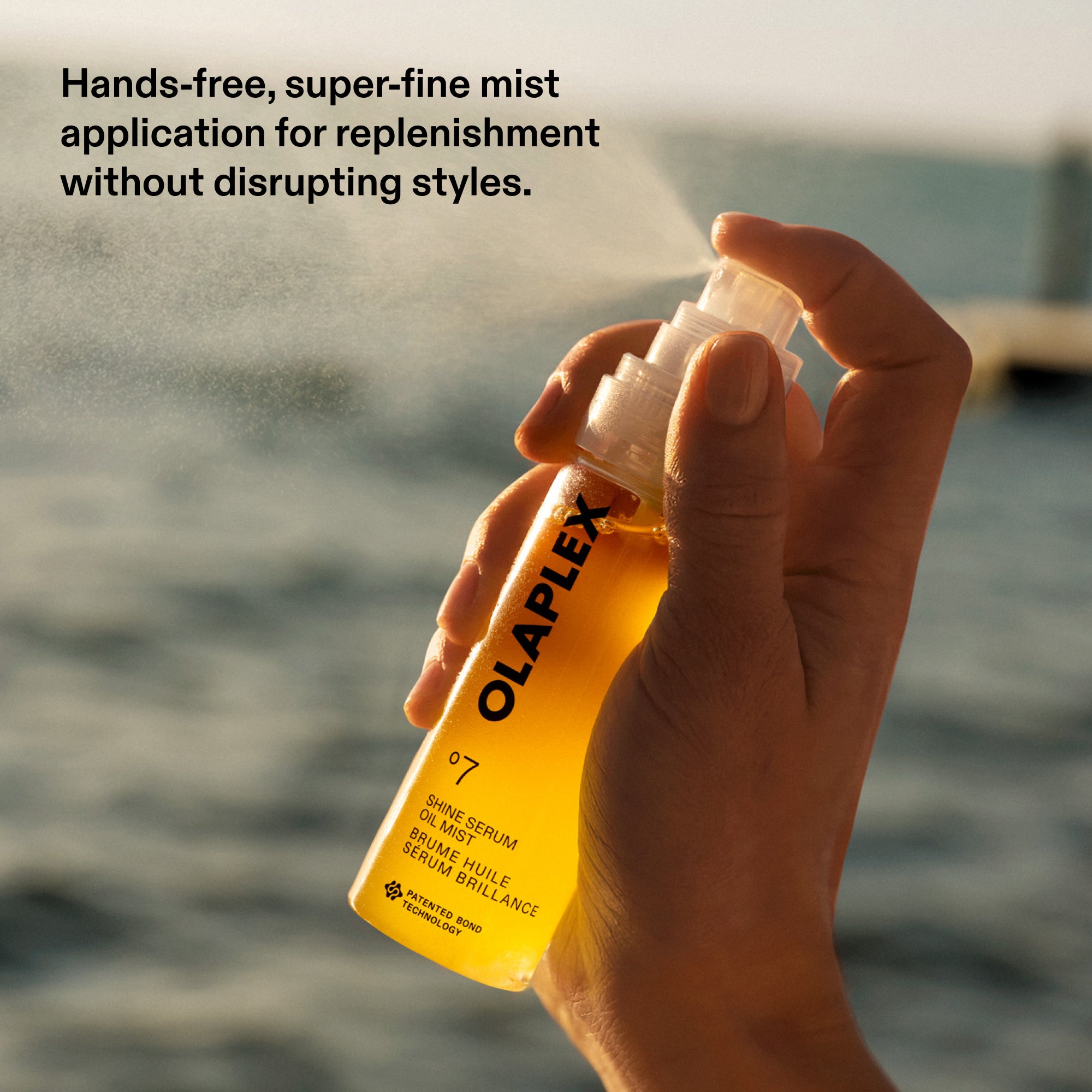 A close-up shot of a hand holding and spraying a bottle of Olaplex No. 7 Shine Serum Oil Mist, releasing a super-fine mist into the air. The golden-yellow liquid is visible inside the clear bottle. In the background, a body of water is softly blurred under bright sunlight. Text in the top-left corner reads: "Hands-free, super-fine mist application for replenishment without disrupting styles.